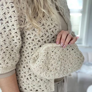 Op de afbeelding: Een beige gehaakt vest en bijpassende clutch. Het vest heeft een bloemenpatroon, korte mouwen en een knoopsluiting. De clutch, in de hand gehouden, heeft een vergelijkbaar bloemenontwerp. De outfit is neutraal van kleur.