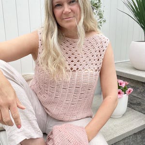 May include: A pale pink, sleeveless crochet top with an openwork yoke and a ribbed body. The top is worn with light-coloured trousers and a matching pink crocheted blanket. The overall aesthetic is soft and feminine, with a focus on handmade details.