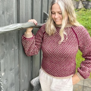 May include: A burgundy and cream-colored knit sweater with a textured pattern. The sweater has long sleeves and a crew neck with cream-colored trim. The person is wearing white pants. The sweater is a cozy and stylish garment.