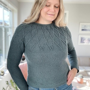 Könnte beinhalten: Ein teal-farbener Langarmpullover mit Rundhalsausschnitt und einem strukturierten, rautenförmigen Muster auf der oberen Brust. Der Pullover wird mit einer blauen Jeans kombiniert. Die Frau auf dem Bild trägt den Pullover.