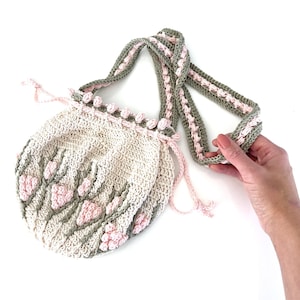 May include: A crocheted bag with a pink and green floral design. The bag has a drawstring closure and a long strap.