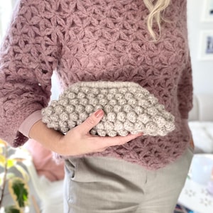 Puede incluir: Un suéter de manga larga color malva con un estampado floral. Un clutch texturizado de color beige claro se sostiene frente al suéter. El clutch tiene una textura redondeada y con bultos. El suéter y el clutch parecen hechos a mano.