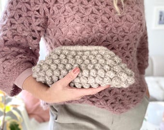 May include: A mauve-colored, long-sleeved sweater with a floral pattern. A light beige, textured clutch is held in front of the sweater. The clutch has a rounded, bumpy texture. The sweater and clutch appear to be handmade.