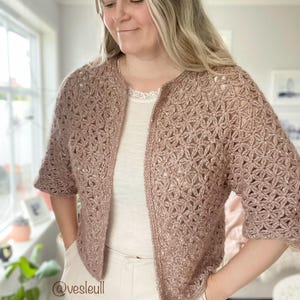 Op de afbeelding: Een lichtroze gehaakte cardigan met een delicaat bloemenpatroon. De cardigan heeft korte mouwen en een licht getailleerde lengte. De cardigan is open aan de voorkant en heeft een eenvoudig, elegant ontwerp.