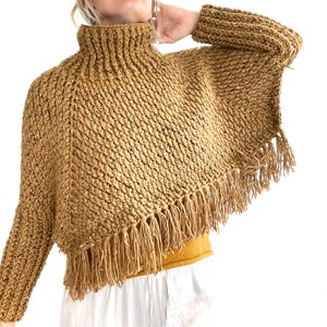 May include: A brown crocheted poncho with long sleeves and a high neck. The poncho has a fringe trim around the bottom edge.