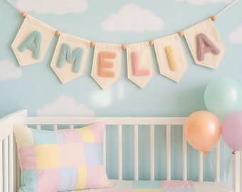 May include: A decorative banner featuring the name AMELIA in pastel-colored, textured, three-dimensional letters on cream-colored fabric pennants, strung together with wooden beads, accompanied by a pastel patchwork pillow and balloons.