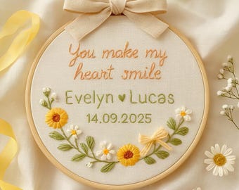 May include: A circular wooden embroidery hoop containing cream fabric with the embroidered text "You make my heart smile", the names "Evelyn" and "Lucas" separated by a green heart, and the date "14.09.2025". A beige fabric bow sits on top.