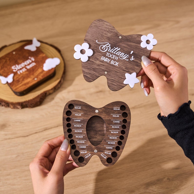 May include: A wooden tooth-shaped tooth fairy box with the name "Brittany's" and "TOOTH FAIRY BOX" in white script. The box has a removable top decorated with white flowers and a star. The bottom section has holes labeled for teeth.