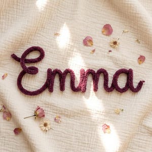 May include: A dark purple, fuzzy, cursive-style wire word sign spelling the name Emma. The letters are formed from a soft, textured yarn-wrapped material, creating a continuous script. Scattered around the word are several dried flower petals.