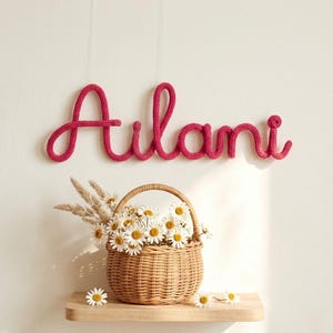 May include: A hot pink knitted wire wall sign spelling the name Ailani in cursive script, positioned above a light brown woven wicker basket filled with white daisy flowers and dried grasses, all resting on a light wooden shelf.