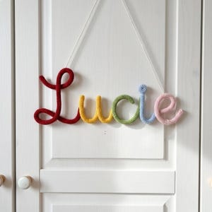 May include: A wall hanging featuring the name Lucie spelled out in cursive, knitted yarn letters. The L is dark red, the u and c are yellow, the i is light green, the dot is light blue, and the e is light pink, suspended by a white string.