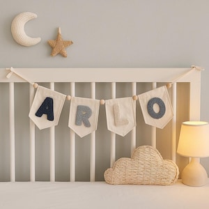 Op de afbeelding: Een slinger voor de kinderkamer met de naam "ARLO" in marineblauwe, lichtblauwe en beige letters op witte vlaggetjes. Een ster en een maan van vlechtwerk hangen boven de slinger. Een wolkvormig kussen en een lamp staan ook in de afbeelding.