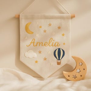Könnte beinhalten: Ein beiges Wandbehang aus Stoff mit dem Namen "Amelia" in goldener Schrift gestickt, zusammen mit einem gelben Halbmond, Sternen, einer Wolke und einem blau-braunen Heißluftballon. Ein hölzerner Halbmond mit Sternaussparungen befindet sich darunter.