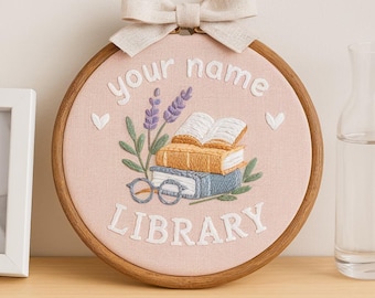 May include: A circular wooden embroidery hoop containing light pink fabric with hand-stitched designs. The embroidery features a stack of three books, a pair of glasses, lavender sprigs, two hearts, and the text "your name" and "LIBRARY". A beige bow is attached to the top.