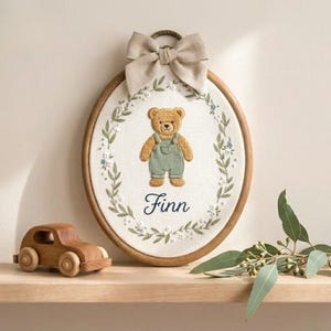 May include: An embroidered oval wall hanging featuring a teddy bear wearing green overalls, surrounded by a floral wreath. The name "Finn" is written in cursive below the bear. A wooden toy car sits on a shelf below the hanging, and a beige bow tops the frame.