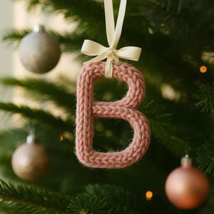 Può includere: Ornamento natalizio a forma di lettera "B" rosa antico, lavorato a maglia, appeso a un nastro color crema legato con un fiocco. L'ornamento è appeso davanti a un albero di Natale con altre palline.