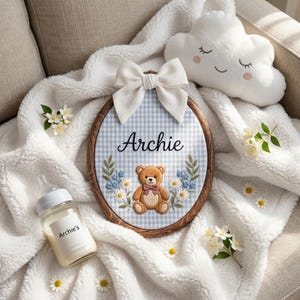 Op de afbeelding: Een ovale houten borduurring met een lichtblauw en wit gingham stoffen achtergrond. De stof is geborduurd met de naam Archie in zwart schrift, een bruine teddybeer met een roze strik en blauwe en witte bloemaccenten.