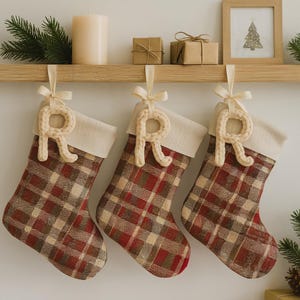 May include: Three Christmas stockings with a red, brown, and tan plaid pattern. Each stocking has a cream-colored cuff and a cream-colored knitted letter "R" hanging from a ribbon. The stockings are hanging on a wooden shelf with other holiday decorations.