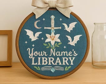 May include: A circular wooden embroidery hoop containing teal fabric with white thread embroidery. The design features a sword, a lily, three bats, a crescent moon, and the text "Your Name's LIBRARY" above an open book. A beige fabric bow is attached to the top.