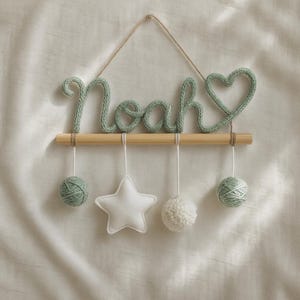 May include: A mint green knitted wall hanging spelling "Noah" with a heart symbol. The name is suspended from a wooden dowel, with hanging yarn balls, a star, and a pom-pom. The background is a light fabric.