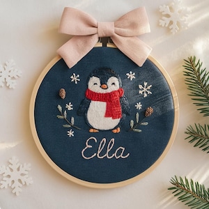 May include: Embroidered Christmas ornament featuring a penguin wearing a red scarf, surrounded by snowflakes and pine sprigs. The name "Ella" is stitched below the penguin. A pink bow tops the wooden hoop.