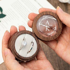 May include: Two wooden ring boxes, one open and one closed, held in hands. The open box displays two rings on a white cushion. The closed box has a clear top with engraved initials and a date. The boxes are brown with a circular design.