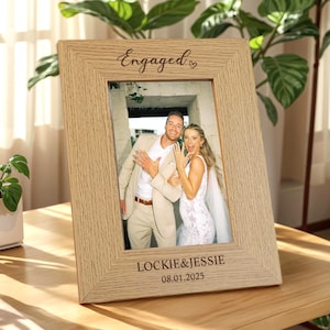 May include: A light-colored wooden picture frame with the word "Engaged" and a heart symbol engraved at the top. The frame holds a photo of a couple. The names "LOCKIE & JESSIE" and the date "08.01.2025" are engraved at the bottom.