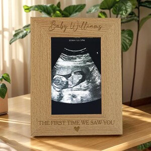 May include: A light oak wood picture frame featuring the engraved text Baby Williams at the top and THE FIRST TIME WE SAW YOU with a small heart icon at the bottom, containing a black and white ultrasound scan of a fetus.