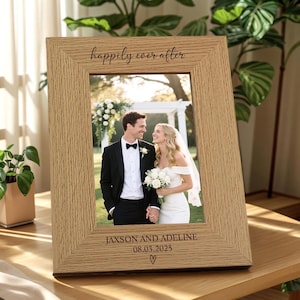 Puede incluir: Un marco de fotos de madera clara con la frase "happily ever after" en la parte superior. Contiene una foto de boda de una pareja. Los nombres "Jaxson and Adeline", la fecha "08.03.2025" y un corazón están grabados en la parte inferior. El marco está sobre una mesa de madera.