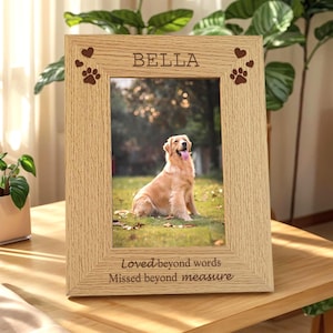 Op de afbeelding: Een lichtgekleurde houten fotolijst met een foto van een golden retriever hond. De lijst heeft de naam "BELLA" bovenaan, pootafdrukken en harten, en de tekst "Loved beyond words, Missed beyond measure" onderaan.