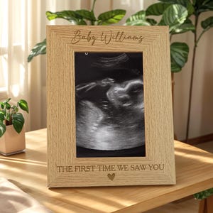 Könnte beinhalten: Ein hellfarbiger Holzbilderrahmen mit dem Text "Baby Williams" und "THE FIRST TIME WE SAW YOU" eingraviert. Der Rahmen enthält ein Schwarz-Weiß-Ultraschallbild eines Babys. Unter dem Text ist ein kleines Herz eingraviert.
