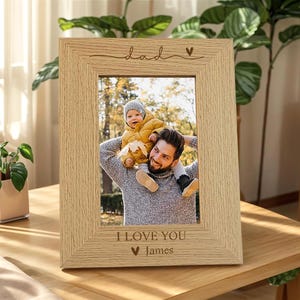 May include: A light-colored wooden picture frame with the word "dad" and a heart symbol engraved at the top. The frame holds a photograph of a man and a child. The bottom of the frame reads "I LOVE YOU" and the name "James".