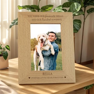 Puede incluir: Un marco de fotos de madera de color claro con el texto "YOU WERE OUR favourite HELLO...AND OUR hardest GOODBYE" y "FOREVER LOVED NEVER FORGOTTEN." El marco contiene una foto de una persona sosteniendo un perro. El nombre del perro, BELLA, también está en el marco.