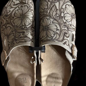 May include: Tan slip-on sandals with a floral embossed design. The sandals have a buckle closure and are size 9. The footbed is tan, and the floral design is a darker brown. The sandals are displayed on a black background.