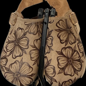 May include: A pair of tan slip-on clogs featuring a dark brown floral line art pattern. Each shoe has a gold-tone metal buckle on the side and a tan footbed with the text Time and Tru size 7 printed on the heel. The clogs are held together by a black plastic hanger.