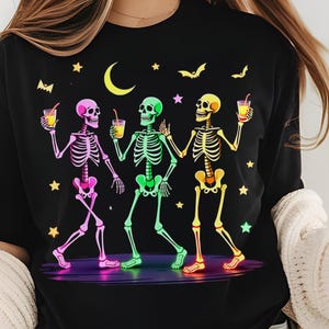 Puede incluir: Camiseta negra con un gráfico colorido de tres esqueletos bailando y sosteniendo bebidas. Los esqueletos están delineados en rosa, verde y amarillo, con un escenario púrpura, estrellas, murciélagos y una luna creciente.
