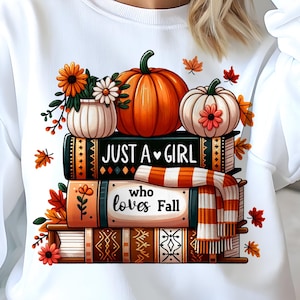 Può includere: Felpa bianca con grafica a tema autunnale. Il design include zucche, fiori e la scritta "Just a Girl who loves Fall" su una pila di libri. Foglie autunnali e una sciarpa completano il tema stagionale.