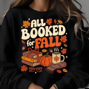 May include: Black sweatshirt with a fall-themed graphic. The design features the words 'All Booked for Fall' in white and orange, along with illustrations of books, a pumpkin, a coffee mug with a leaf, and autumn leaves. The overall aesthetic is cozy and autumnal.