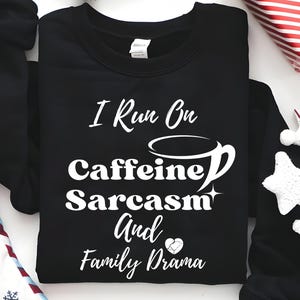 Puede incluir: Sudadera negra con texto blanco "I Run On Caffeine, Sarcasm, And Family Drama". La imagen incluye decoraciones festivas como un regalo envuelto y adornos en forma de estrella.