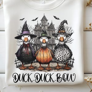 May include: White sweatshirt featuring a Halloween-themed graphic. The graphic depicts three ducks dressed as witches in front of a haunted house, with bats flying above. The text "DUCK. DUCK. BOW" is printed below the ducks.