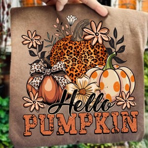 Peut inclure: T-shirt marron avec un motif d'automne. Le motif comprend trois citrouilles, dont une avec un imprimé léopard, et le texte "Hello PUMPKIN" en orange. Des éléments floraux et un nœud complètent le look festif.