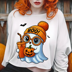 May include: White long-sleeve shirt featuring a cartoon ghost wearing an orange beanie with the word "BOO!" and glasses. The ghost holds a pumpkin and a drink. Two black bats are flying in the background.