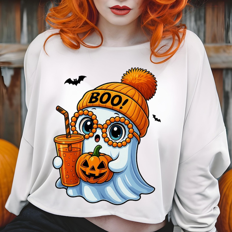 May include: White long-sleeve shirt featuring a cartoon ghost wearing an orange beanie with the word "BOO!" and glasses. The ghost holds a pumpkin and a drink. Two black bats are flying in the background.