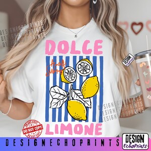 Puede incluir: Camiseta blanca con un dise&ntilde;o gr&aacute;fico que presenta la palabra "DOLCE" en rosa, "Amalfi Summer" en naranja y "LIMONE" en azul. El dise&ntilde;o incluye ilustraciones de limones y rayas verticales azules.