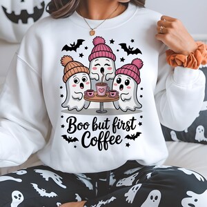 May include: White sweatshirt with a Halloween-themed graphic. The design features three ghost characters wearing pink and brown beanies, gathered around a small table with coffee cups. The text "Boo but first Coffee" is also included.