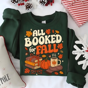 Puede incluir: Una sudadera verde oscuro con el texto "ALL BOOKED for FALL" en crema y naranja. El dise&ntilde;o incluye ilustraciones de libros, una calabaza, una taza y hojas de oto&ntilde;o. Un art&iacute;culo con tem&aacute;tica oto&ntilde;al.