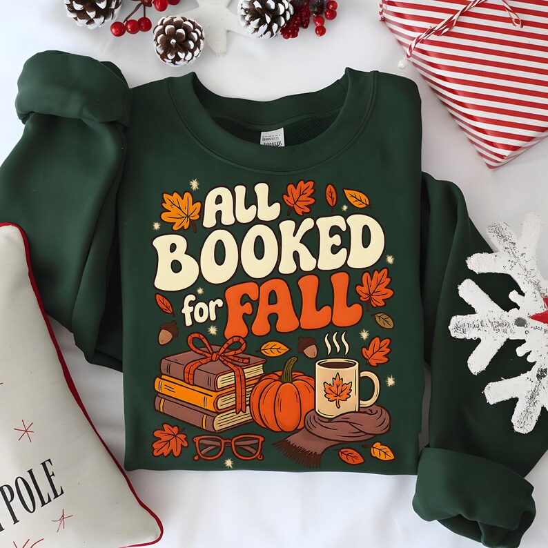 Puede incluir: Una sudadera verde oscuro con el texto "ALL BOOKED for FALL" en crema y naranja. El dise&ntilde;o incluye ilustraciones de libros, una calabaza, una taza y hojas de oto&ntilde;o. Un art&iacute;culo con tem&aacute;tica oto&ntilde;al.