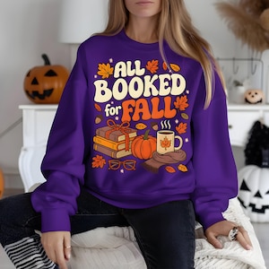 Puede incluir: Una sudadera morada con el texto "ALL BOOKED for FALL" en crema y naranja. El dise&ntilde;o incluye libros, una calabaza, una taza y hojas de oto&ntilde;o. Una sudadera con tem&aacute;tica oto&ntilde;al.