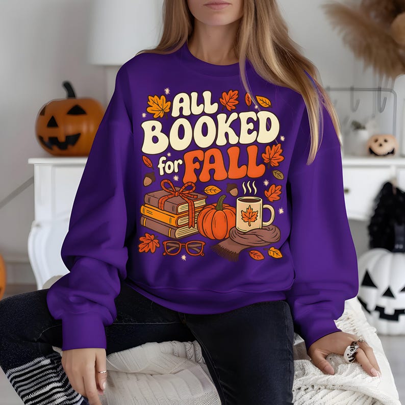 Puede incluir: Una sudadera morada con el texto "ALL BOOKED for FALL" en crema y naranja. El dise&ntilde;o incluye libros, una calabaza, una taza y hojas de oto&ntilde;o. Una sudadera con tem&aacute;tica oto&ntilde;al.