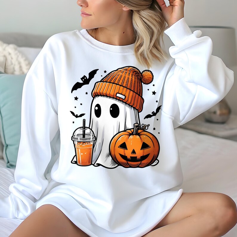 May include: White sweatshirt featuring a cartoon ghost wearing an orange beanie and holding a drink. A carved pumpkin and bats are also in the design. The sweatshirt is a Halloween-themed graphic.